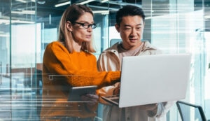 Two colleagues stand together reading something from a laptop screen.