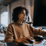 Woman with dark hair in a brown jumper operating a PC using a mouse.
